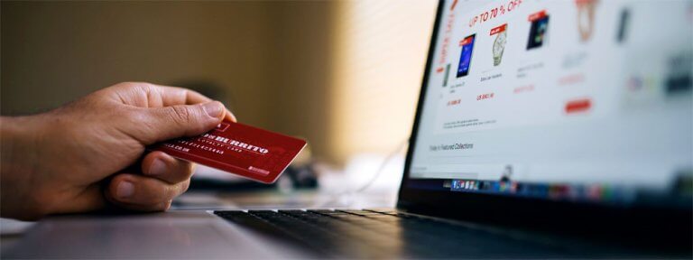 Foto zeigt einen Mann vor dem Laptop, mit einer Kreditkarte in seiner linken Hand. Auf dem Bildschirm ist ein Onlineshop zu sehen